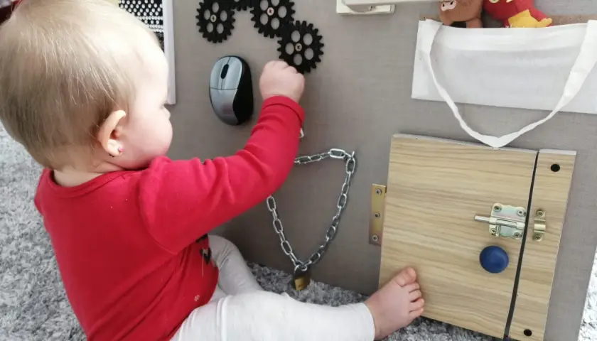 Dziecko bawi się przy tablicy manipulacyjnej diy: zębatki, myszka, zamek, kłódka i pluszaki.