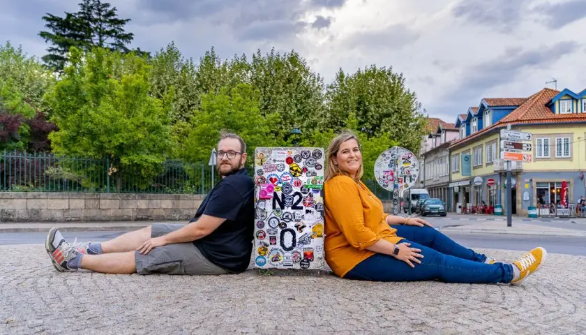 Pareja sentada de espaldas en un poste con pegatinas, celebrando su viaje por la Estrada Nacional 2 en Portugal.