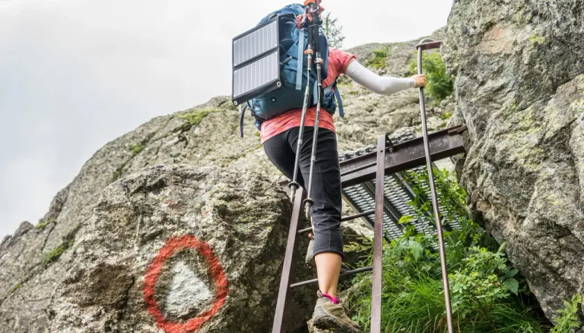 A hiker ascends metal ladders on the Tour du Mont Blanc, using trekking poles and a solar panel on their backpack.