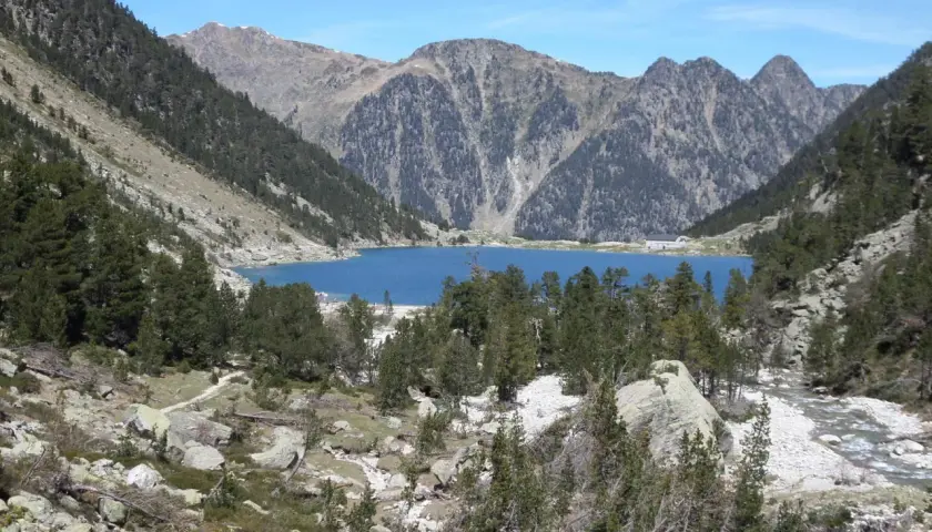 Lago azul rodeado de montañas y pinos, con un pequeño refugio en la orilla. El refugio Oulettes de Gaube es un destino popular para excursionistas.