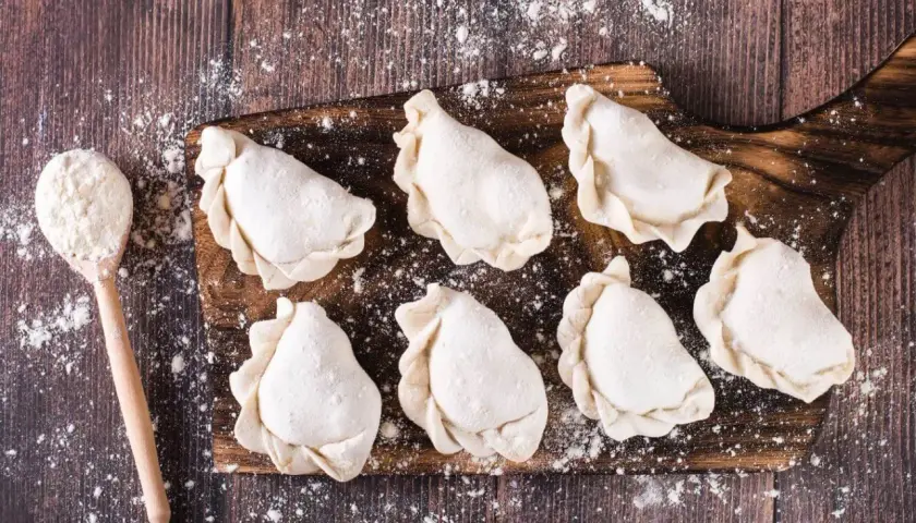 Surowe pierogi na desce, gotowe do przechowywania w lodówce. Obok łyżka z mąką.