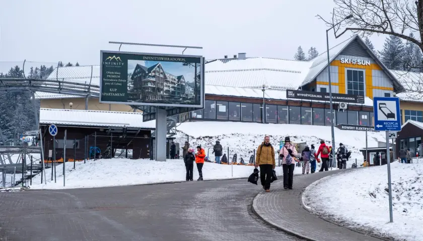 Zimowy dzień w Karpaczu, Świeradowie-Zdroju. Ludzie spacerują przy stoku narciarskim, obok billboardu reklamującego apartamenty.