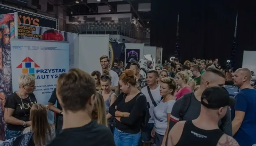 Ludzie na konwencie tatuażu, gdzie widoczny jest symbol autyzmu na banerze "Przystań Autys".