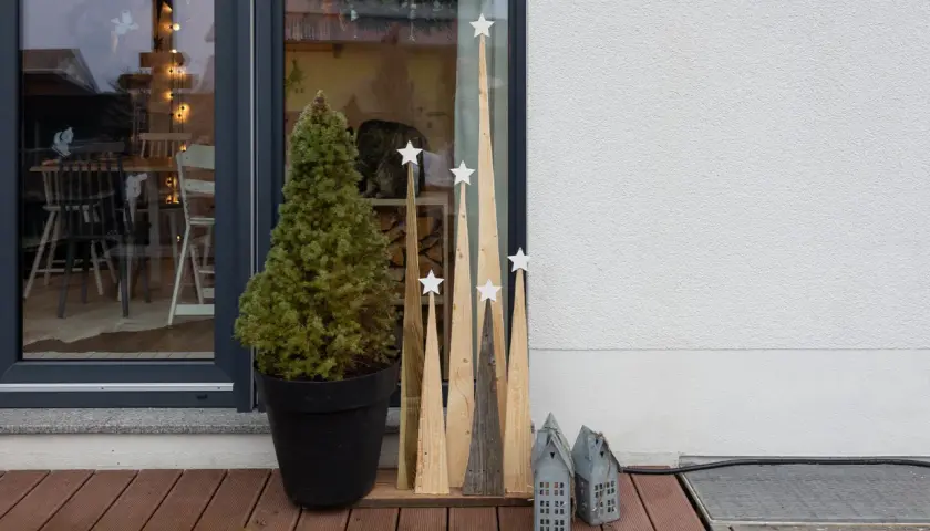 Selbstgemachte Weihnachtsdeko außen: Tannenbaum im Topf, Holz-Tannenbäume mit Sternen und kleine Lichterhäuser auf einer Holzterrasse.