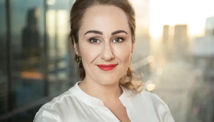 Portretowa fotografia bizneswoman z uśmiechem, w białej bluzce, na tle rozmazanego miasta.