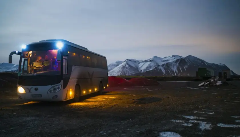 Autobus z zapalonymi światłami na tle ośnieżonych gór o zmierzchu.