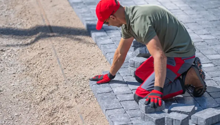 Pracownik w czerwonej czapce układa kostkę brukową. Zastanawiasz się, ile kosztuje ułożenie kostki brukowej? To zależy od wielu czynników.