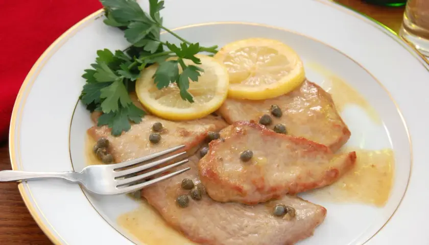 Piccata de veau, tendre escalope de veau sauce citronnée, câpres et persil frais, sur une assiette blanche bordée d'or.