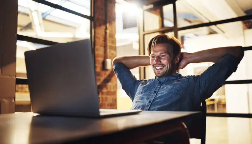 Uśmiechnięty mężczyzna w back office, z rękami za głową, patrzy na laptopa.