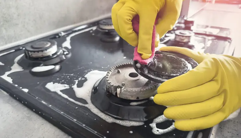Mains jaunes pour nettoyer graisse brûlée gazinière. Une éponge rose frotte une partie du brûleur.