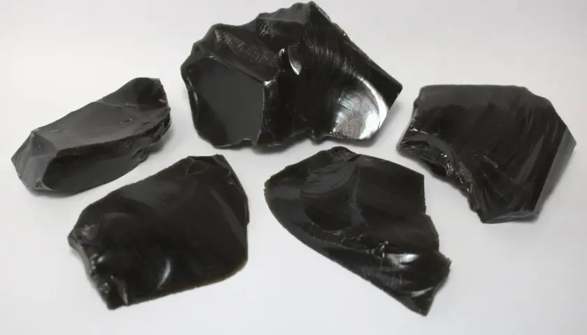 Fünf scharfkantige Stücke glänzenden, schwarzen Obsidiansteins liegen auf weißem Untergrund.