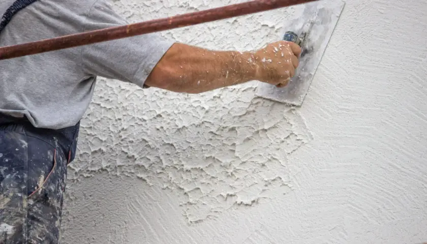 Un operaio applica intonaco rustico con una spatola, creando una texture grezza e irregolare su una parete esterna.
