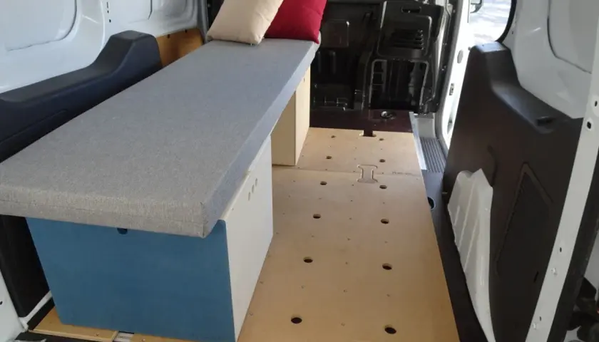 Aménagement intérieur d'un petit fourgon : banquette grise avec coussins, caisson de rangement bleu et blanc, plancher en bois. Prêt pour l'aventure !