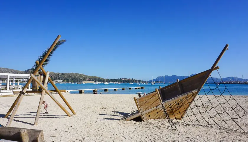 Spielplatz am Strand im mallorcas norden. Ein hölzernes Schiffswrack und eine Kletterstruktur stehen im Sand, Boote liegen im türkisfarbenen Wasser.