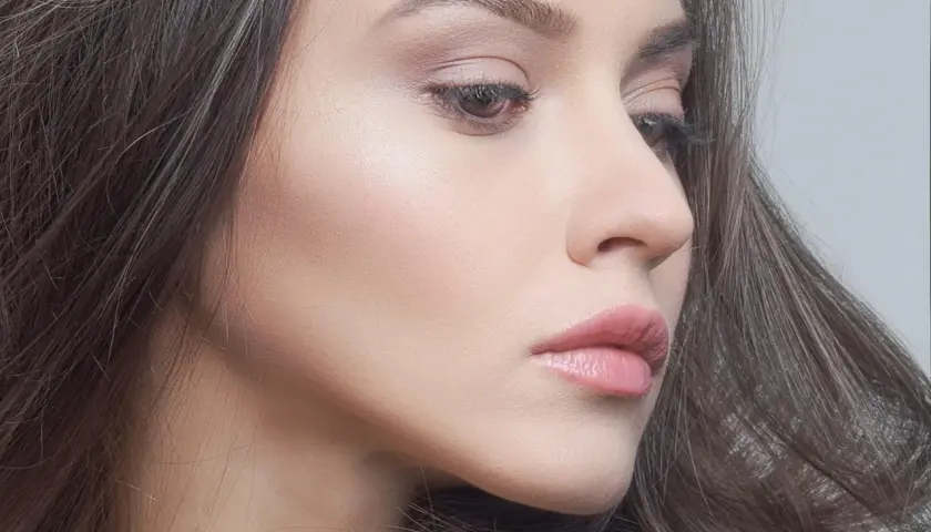 Delikatny profil kobiety z subtelnym makijażem. Zimne kolory podkreślają jej urodę, a włosy tworzą miękką ramę.