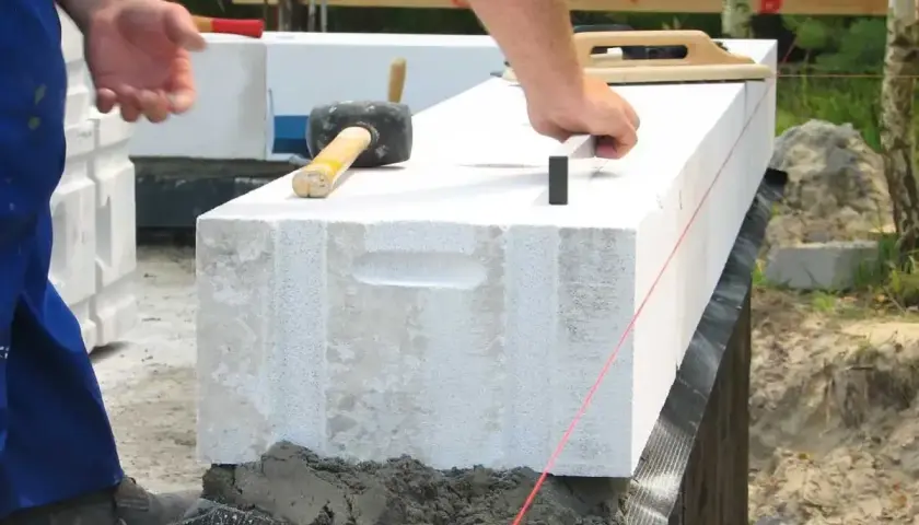 Budowanie ścian z bloczków betonowych z użyciem zaprawy murarskiej. Robotnik układa kolejny bloczek, używając gumowego młotka.