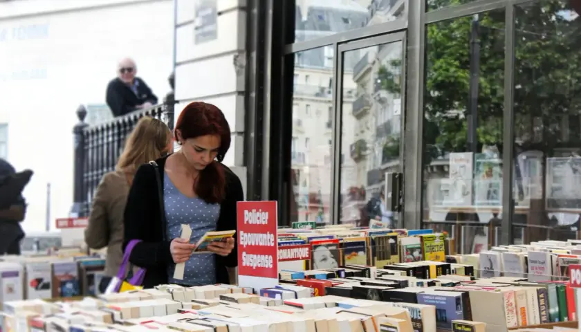 Une jeune femme choisit un livre dans une librairie en plein air, devant une vitrine reflétant des bâtiments parisiens. Une sélection de livres, dont des romans policiers et de suspense, est exposée, rappelant une maison d'édition jeunesse.