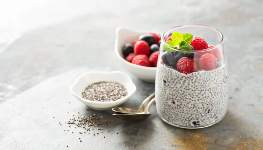 Pudding chia z malinami i borówkami, ozdobiony miętą, obok miseczki z nasionami chia.