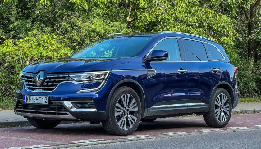 Granatowy Renault Koleos stoi na czerwonej kostce brukowej, otoczony zielenią.