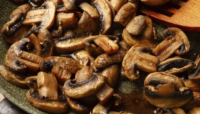 Gebratene Champignons in einer Pfanne, dampfend und glänzend, mit einem hölzernen Kochlöffel.