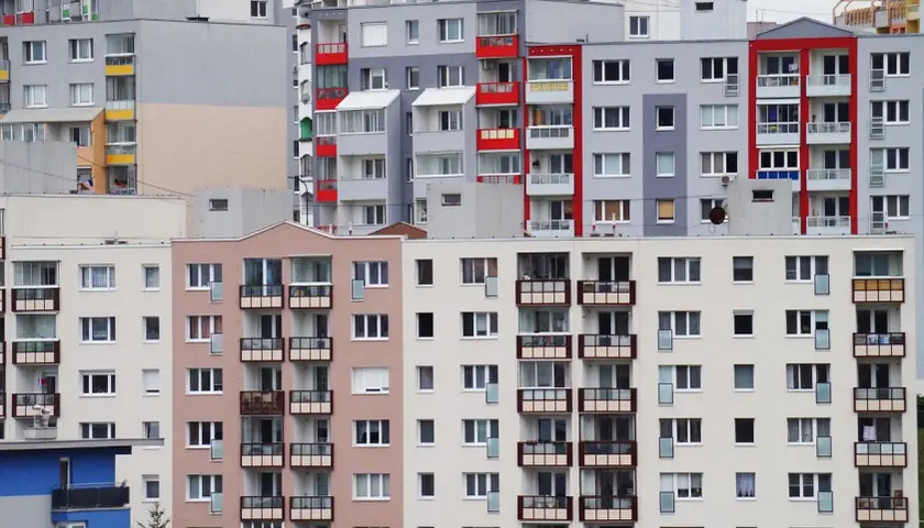 Wielopiętrowe bloki mieszkalne z balkonami. W jednym z mieszkań widoczna jest kartka z napisem "reklamacja z tytułu rękojmi".