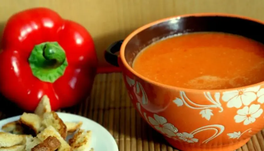 Aksamitny zupa krem z papryki, podana z chrupiącymi grzankami. Obok leży świeża, czerwona papryka.