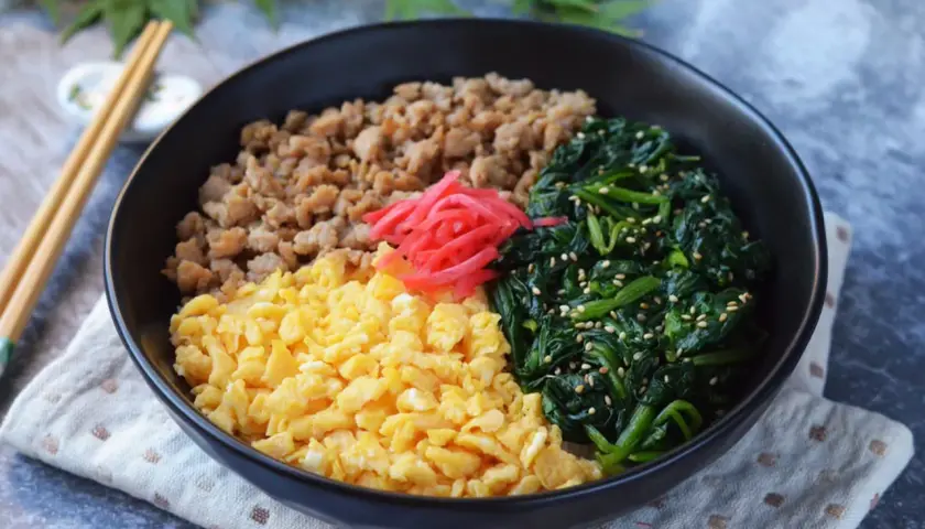 A colorful sanshoku don bowl with scrambled eggs, seasoned ground meat, and spinach, topped with pickled ginger.