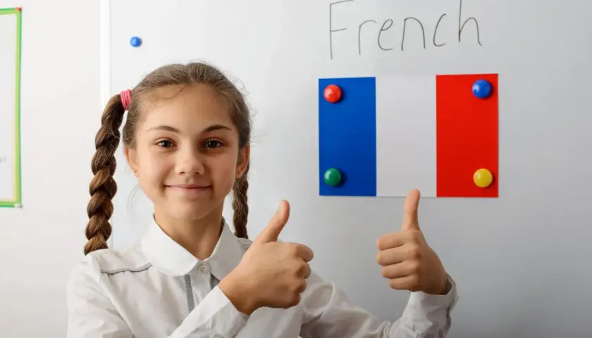 Uśmiechnięta dziewczynka z warkoczami pokazuje kciuki w górę. Na tablicy napis "French" i flaga Francji. Edukacja we Francji ciekawostki.