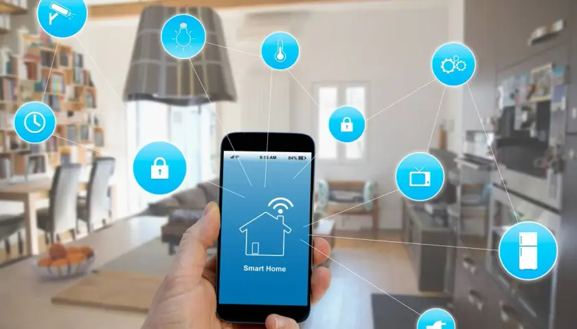 Mano sujetando smartphone con app "Smart Home" para control de instalación domótica. Iconos de seguridad, luz, temperatura, TV, nevera y enchufe conectados.