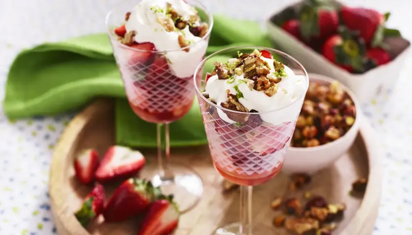Två glas med marinerade jordgubbar, vispgrädde och nötter. En skål med jordgubbar och en skål med nötter syns också.