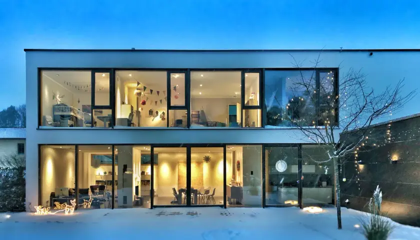 Casa moderna con grandes ventanales iluminados, mostrando interiores acogedores. La nieve cubre el exterior, y luces navideñas decoran un árbol. ¡2500 lumens cuanto ilumina!