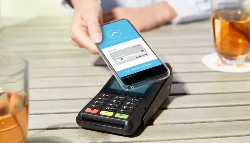 Mit handy bezahlen: Eine Hand hält ein Smartphone über ein Kassenterminal, um kontaktlos zu bezahlen.