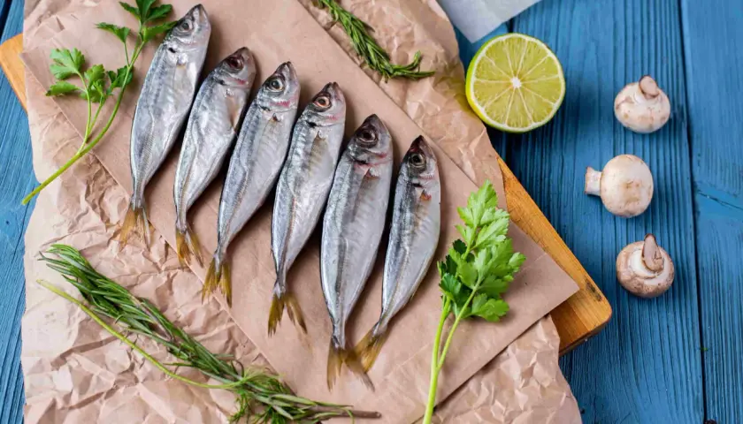 Seis jureles pescado frescos, listos para cocinar, acompañados de perejil, romero, limón y champiñones sobre papel de estraza y tabla de madera.