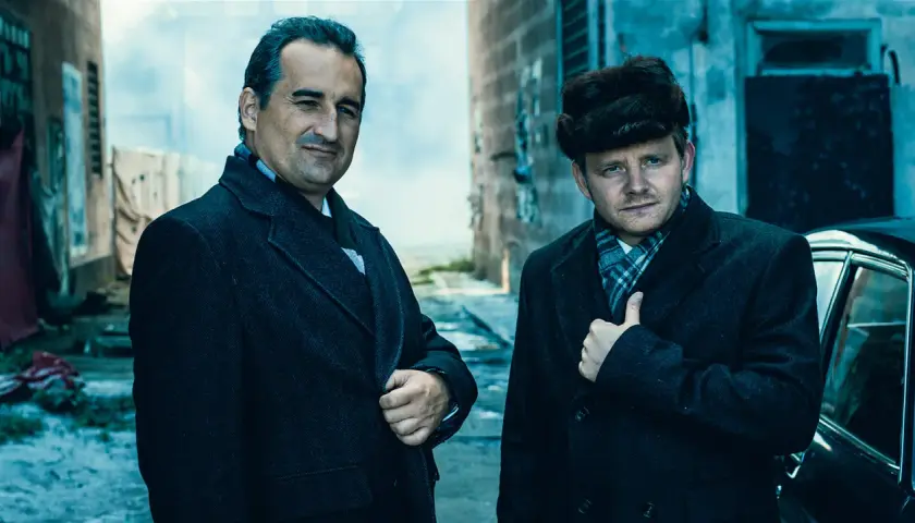 Obsada filmu "Gierek": dwaj mężczyźni w płaszczach i szalikach, jeden w futrzanej czapce, stoją obok czarnego samochodu.