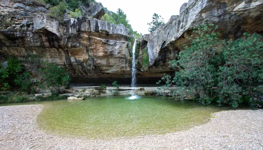 Cascada cayendo en **charcos de quesa** de agua cristalina, rodeada de rocas y vegetación exuberante.