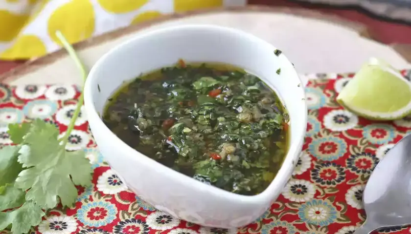 Un tazón blanco con una deliciosa **receta de chimichurri** casera, adornado con cilantro fresco y una rodaja de limón.