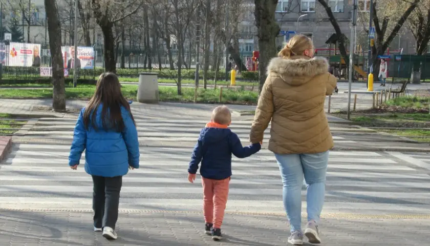 Rodzina przechodzi przez pasy. Kobieta trzyma za rękę małego chłopca, a obok idzie starsza dziewczynka. Szczególna ostrożność kiedy przechodzimy przez jezdnię.