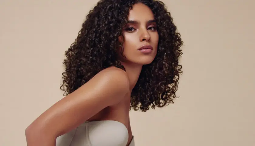 Mujer con semirecogido pelo rizado, luciendo un top beige. Su cabello oscuro y voluminoso cae en cascada sobre sus hombros.