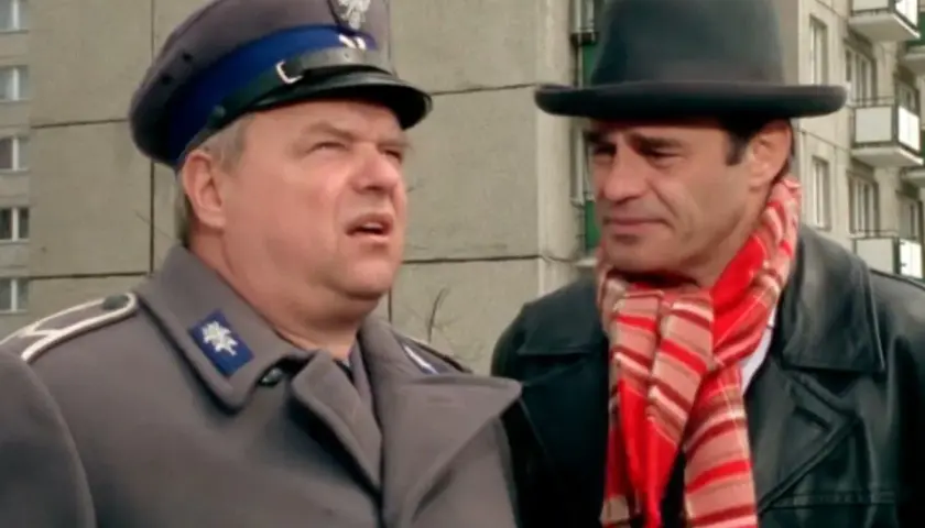 Dwóch mężczyzn, jeden w mundurze milicyjnym, drugi w kapeluszu i szaliku, w kadrze z kultowych bareja filmów.