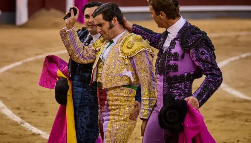 Tres toreros con trajes de luces, uno con coleta de torero, son felicitados en la arena.