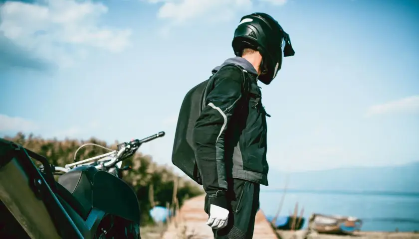 Motociclista con casco negro, chaqueta y guantes blancos, junto a su moto. El tejido de poliéster 600d vs Cordura es ideal para su aventura.