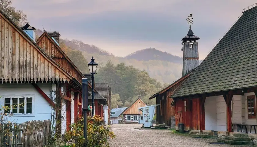 Sanok skansen: urokliwa, brukowana uliczka z drewnianymi domami i wieżą z wiatrowskazem, otoczona mglistymi wzgórzami.