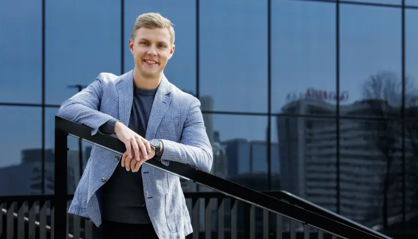 Uśmiechnięty mężczyzna w marynarce w kratę podczas sesji biznesowej w plenerze, oparty o balustradę na tle nowoczesnego budynku.