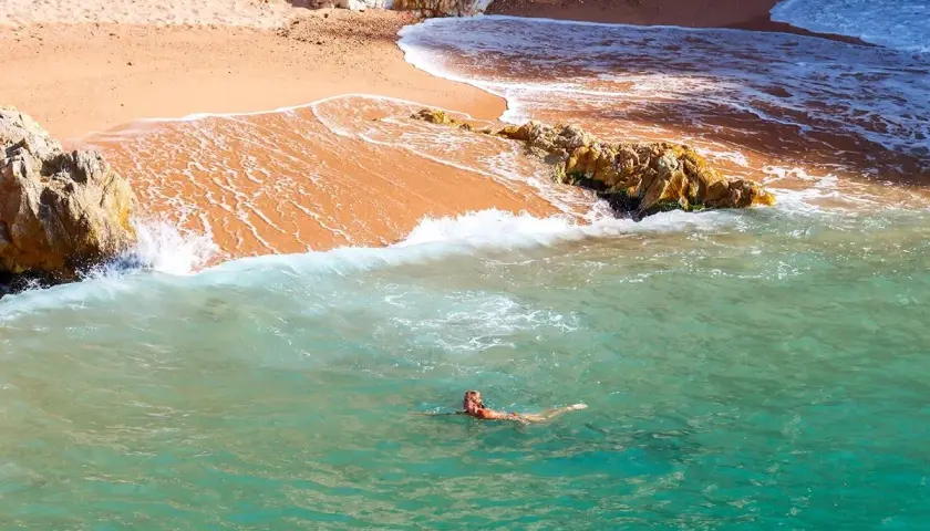 Eine Frau schwimmt im türkisfarbenen Wasser vor einem goldenen costa brava strand, umgeben von Felsen.