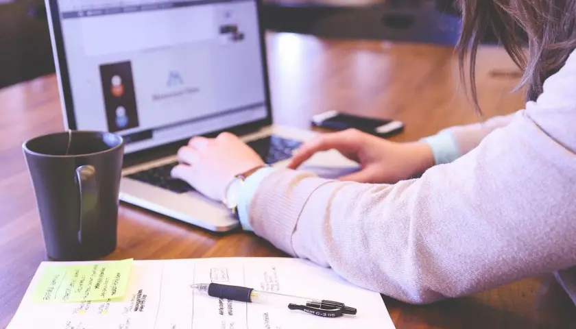Kobieta pracuje przy laptopie. Na biurku leżą notatki, długopis i kubek. Dobre natężenie oświetlenia na stanowisku pracy sprzyja koncentracji.