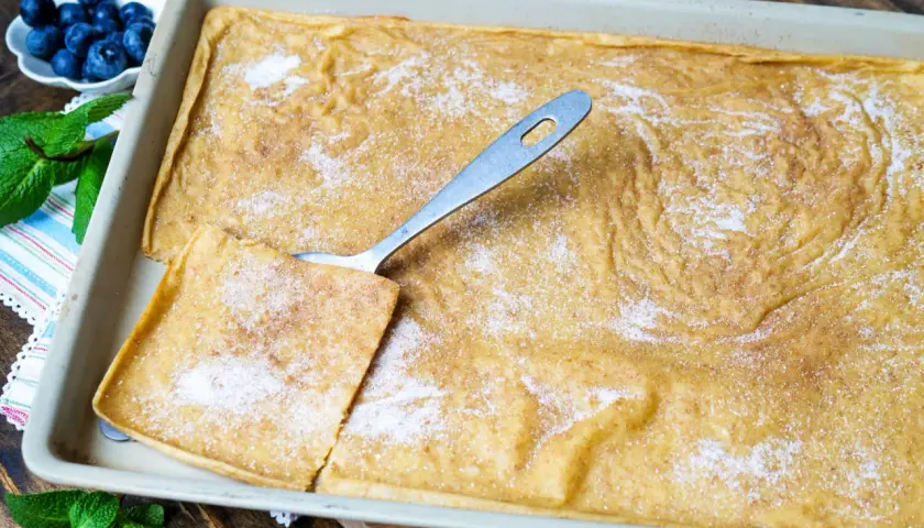 Ein frisch gebackener Ofenpfannkuchen, bestreut mit Zucker und Zimt, wartet darauf, serviert zu werden. Ein Stück ist bereits herausgeschnitten.