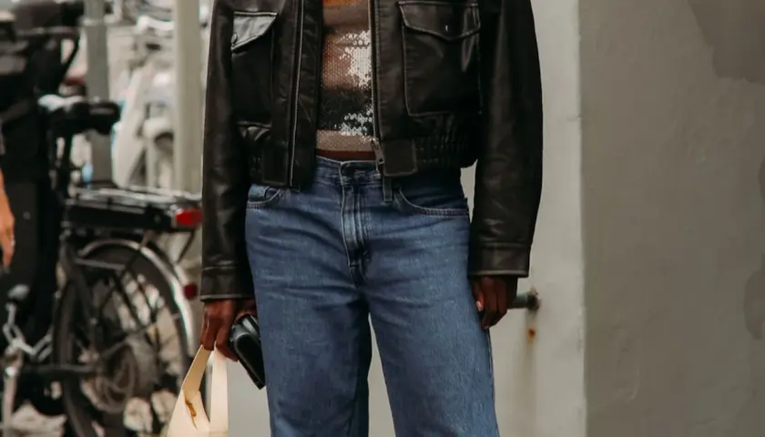 Personne portant une veste en cuir noire, un haut à paillettes et un jean bleu, tenant un sac beige.