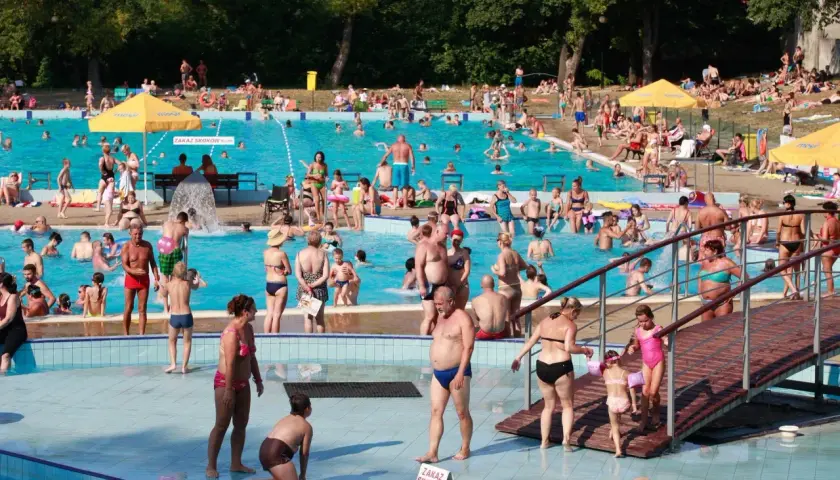 Tłum ludzi korzysta z basenu w upalny dzień. Wszyscy ubrani są w stroje kąpielowe, niektórzy w koszulki na basen, by chronić się przed słońcem.