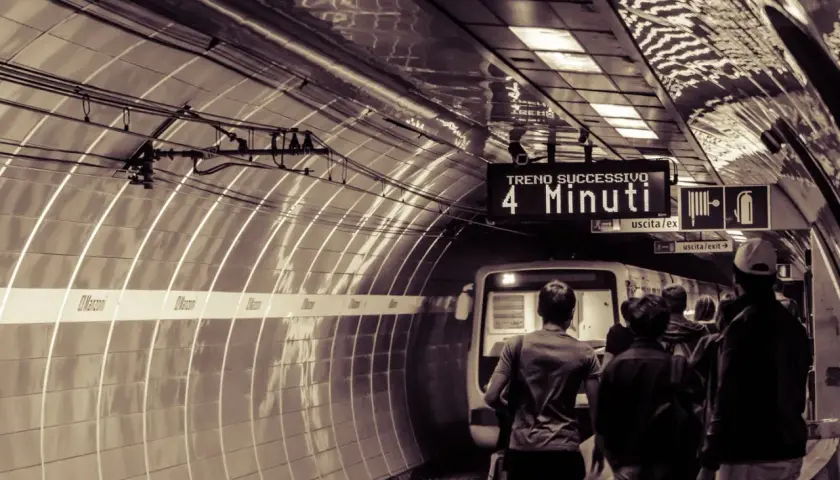 Persone attendono il treno sulla metro linea b. Un cartello indica "Treno successivo: 4 minuti".