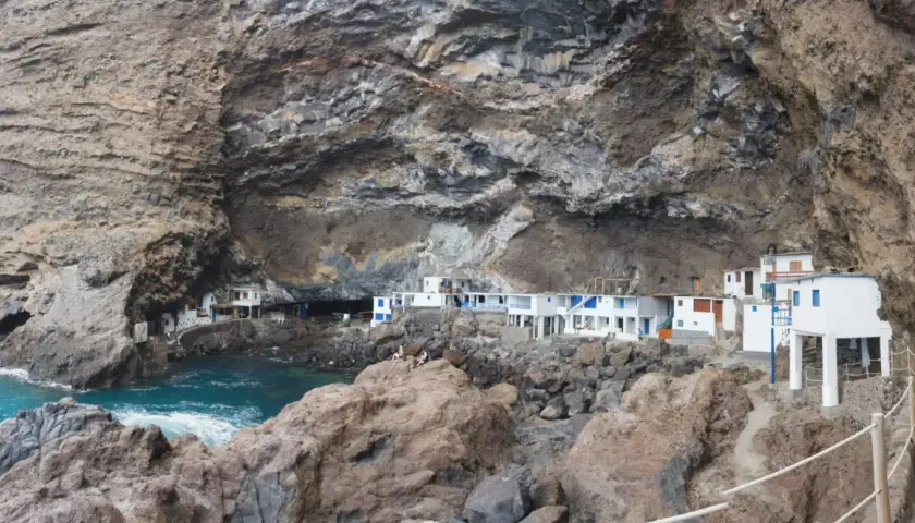 Casas blancas con detalles azules se asoman desde una cueva en los poris de Candelaria, La Palma. El mar turquesa rompe contra las rocas.